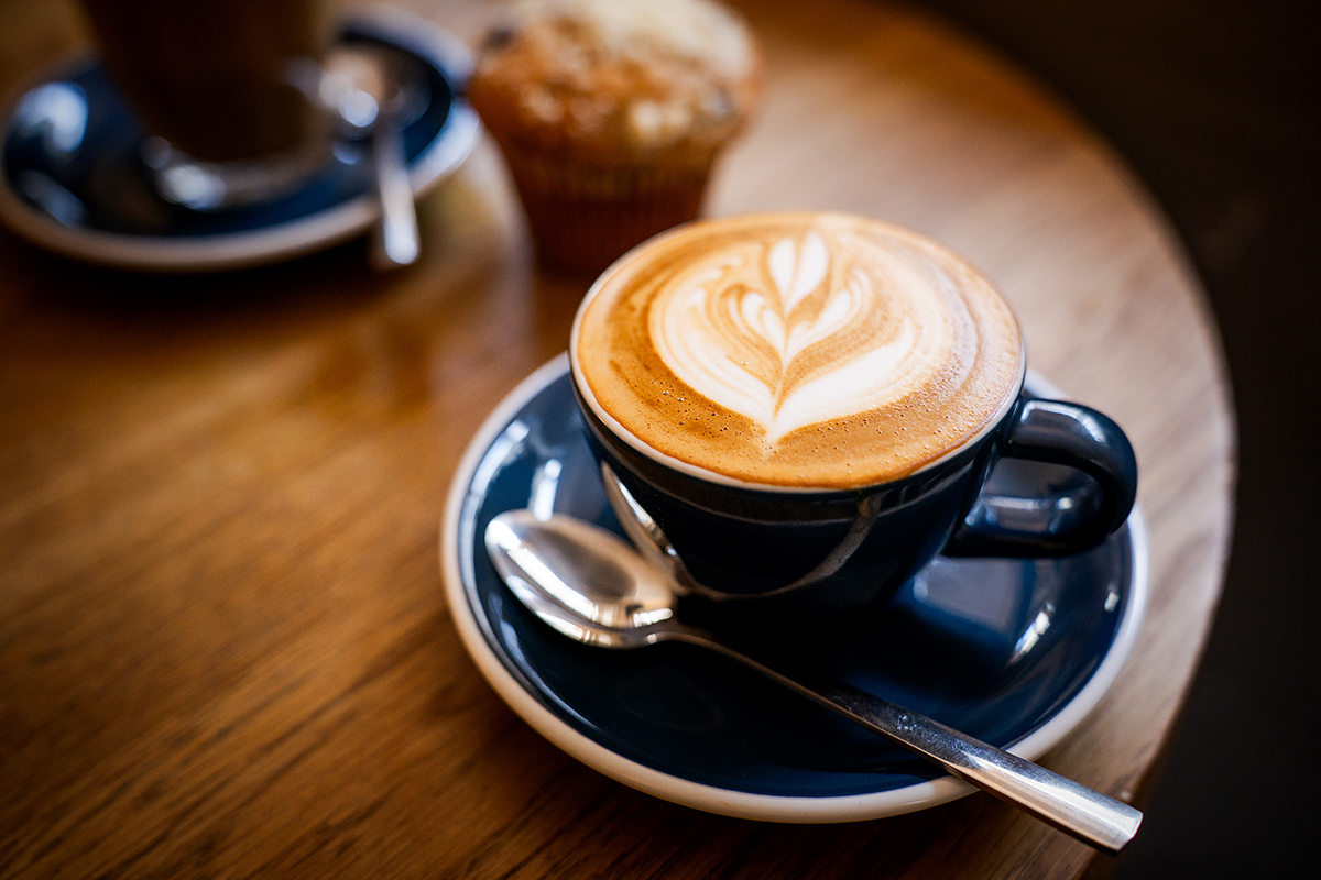 Ein Cappuccino mit Latte-Art in einer dunklen Tasse mit Untertasse auf einem Holztisch, dazu ein Löffel und ein Muffin im Hintergrund.