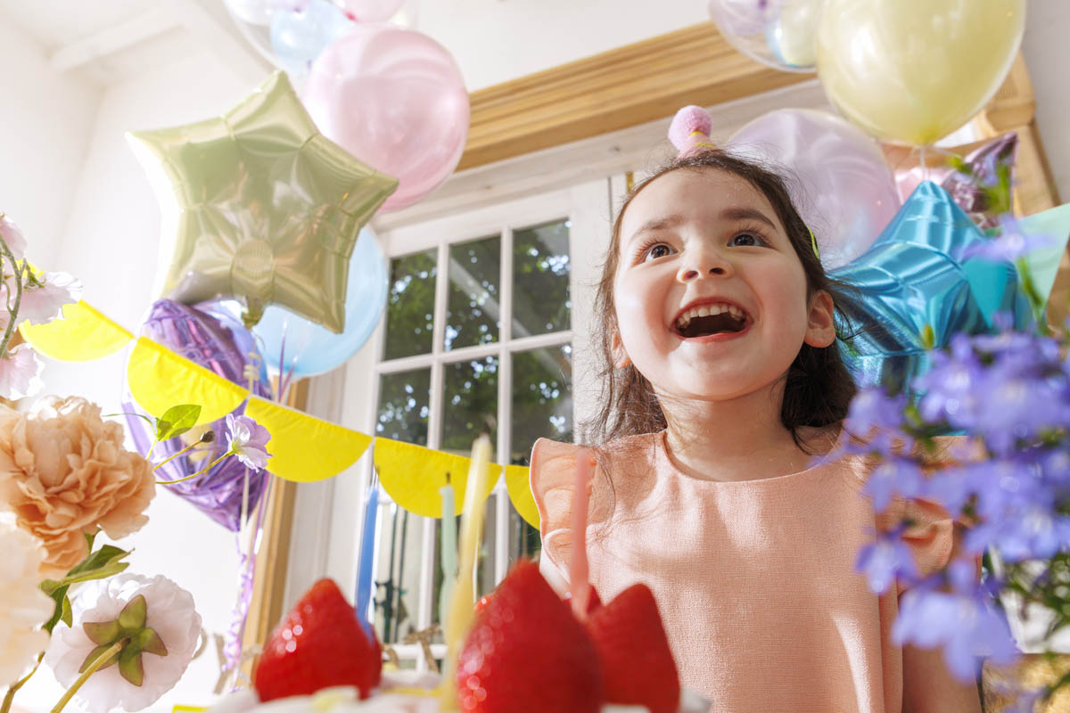 A joyful girl at a vibrant party with colorful balloons, flowers, and a cake in the foreground.