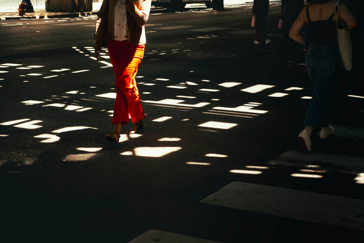 People walking under a bridge creating a patterned shadow on the street, with one person wearing bright red pants.