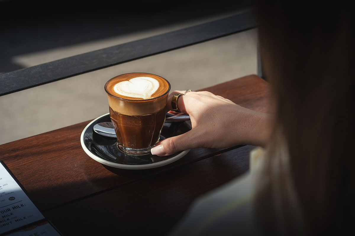 Eine Person hält ein Glas Kaffee mit herzförmiger Latte-Art auf einer Untertasse, das auf einem Holztisch steht.