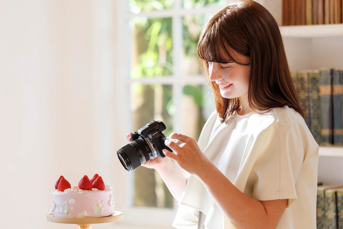 A person takes a photo of a strawberry-topped pink cake with a DSLR camera, in a well-lit room with a large window in the background.