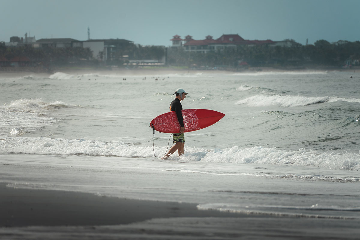 Ein Surfer mit einem roten Surfbrett läuft an einem bewölkten Tag in die Meereswellen, während in der Ferne Gebäude an der Küste zu sehen sind.