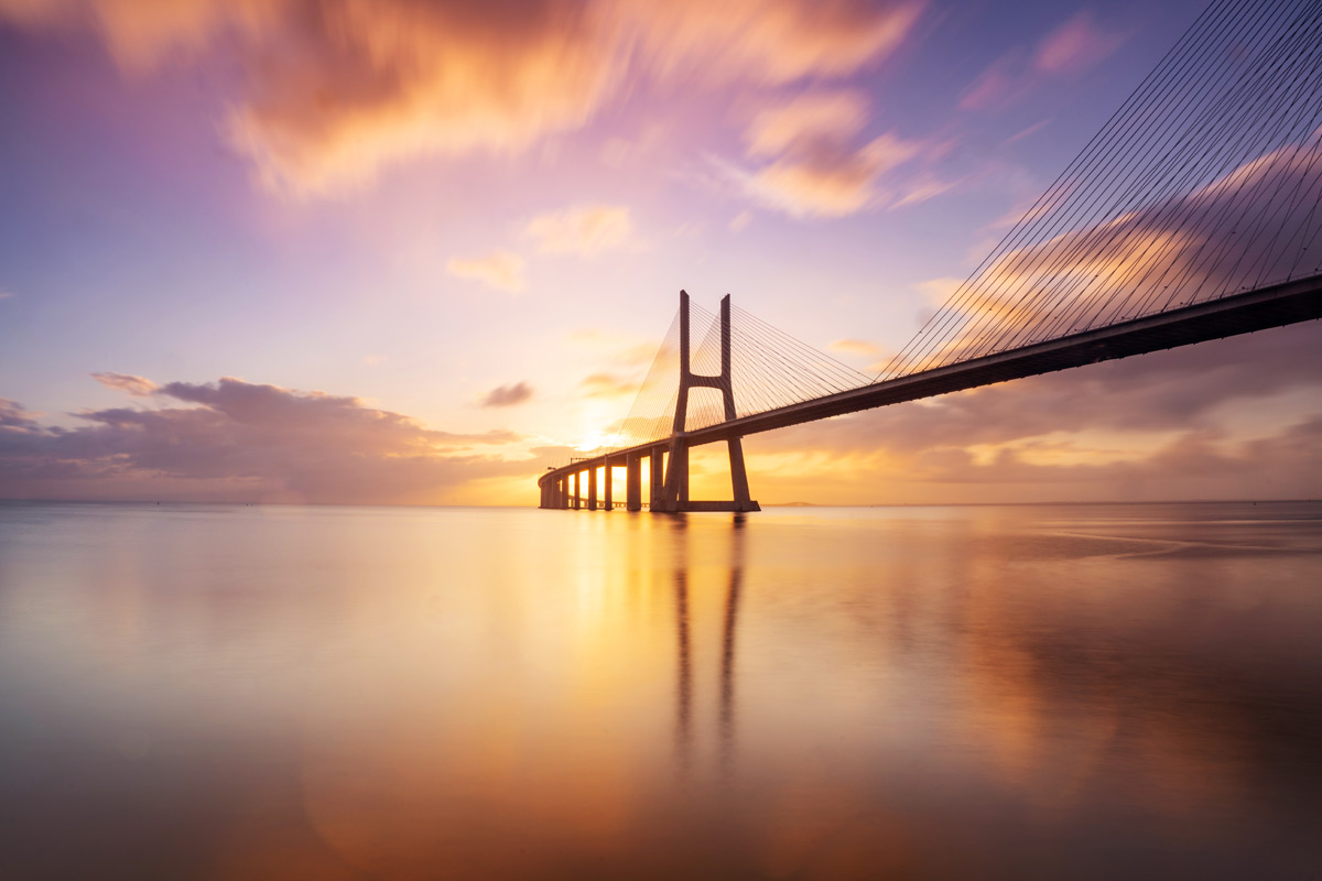 Sonnenuntergangsblick auf eine lange Hängebrücke, die sich über ruhiges Wasser spannt – im Hintergrund ein farbenfroher Himmel mit Wolken.