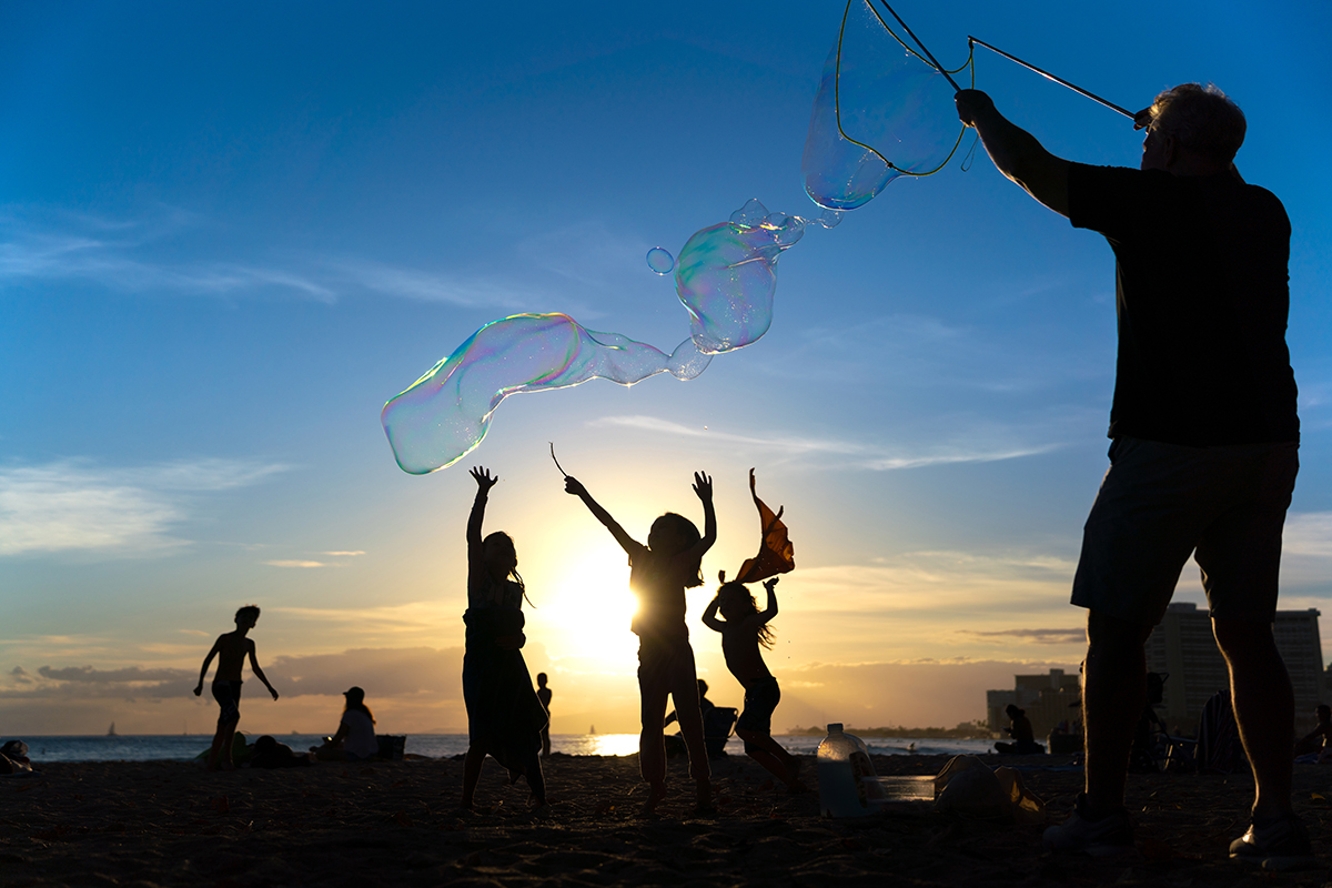 Silhouettes of children reaching for giant bubbles at the beach during sunset, with a person creating bubbles in the foreground.
