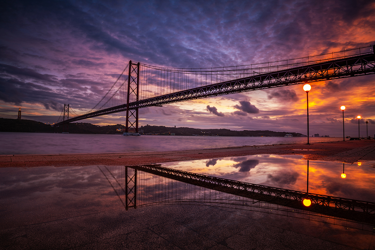The illuminated bridge at sunset is reflected in the water, and the sky is bathed in bright shades of purple and orange. The streetlights shine along the waterfront promenade.