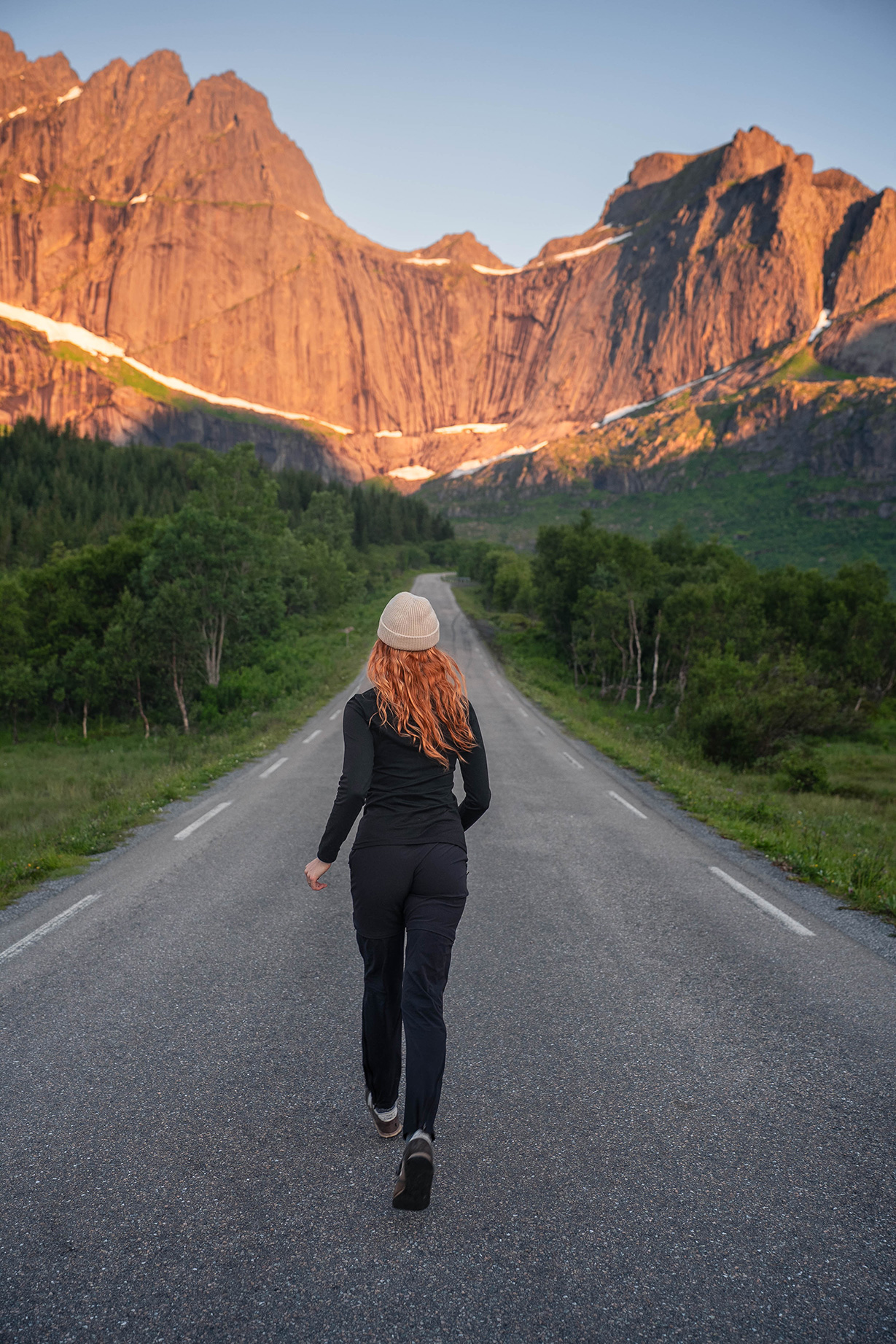 A woman dressed in black clothing and a cap walks down a road toward sunlit mountains surrounded by greenery.
