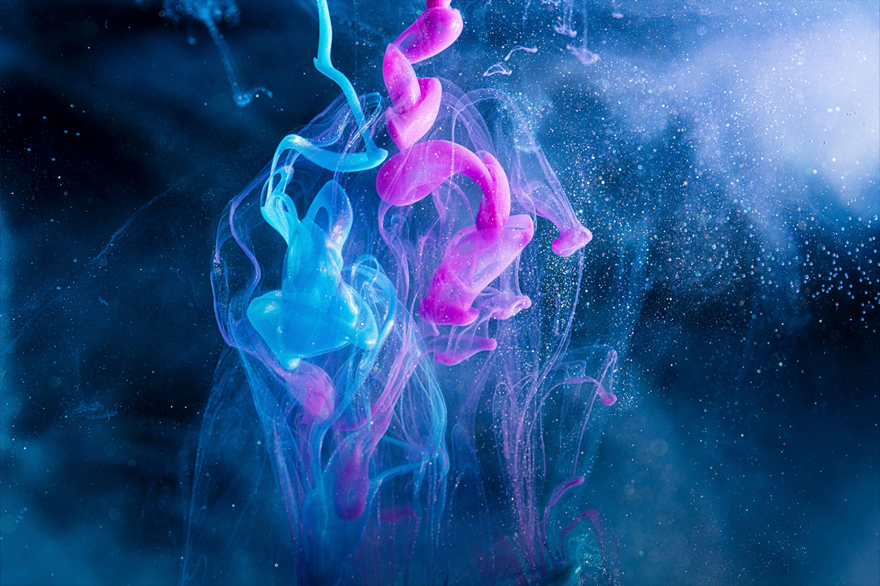 Swirls of pink and blue ink blend and diffuse in water against a soft, misty blue background.