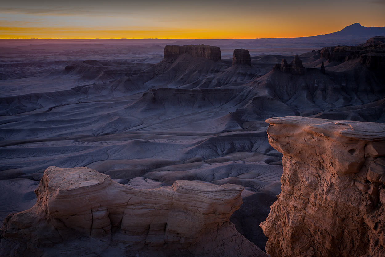 Sunrise over a rocky desert landscape with layered canyons and a distant mountain silhouetted against the colorful horizon.