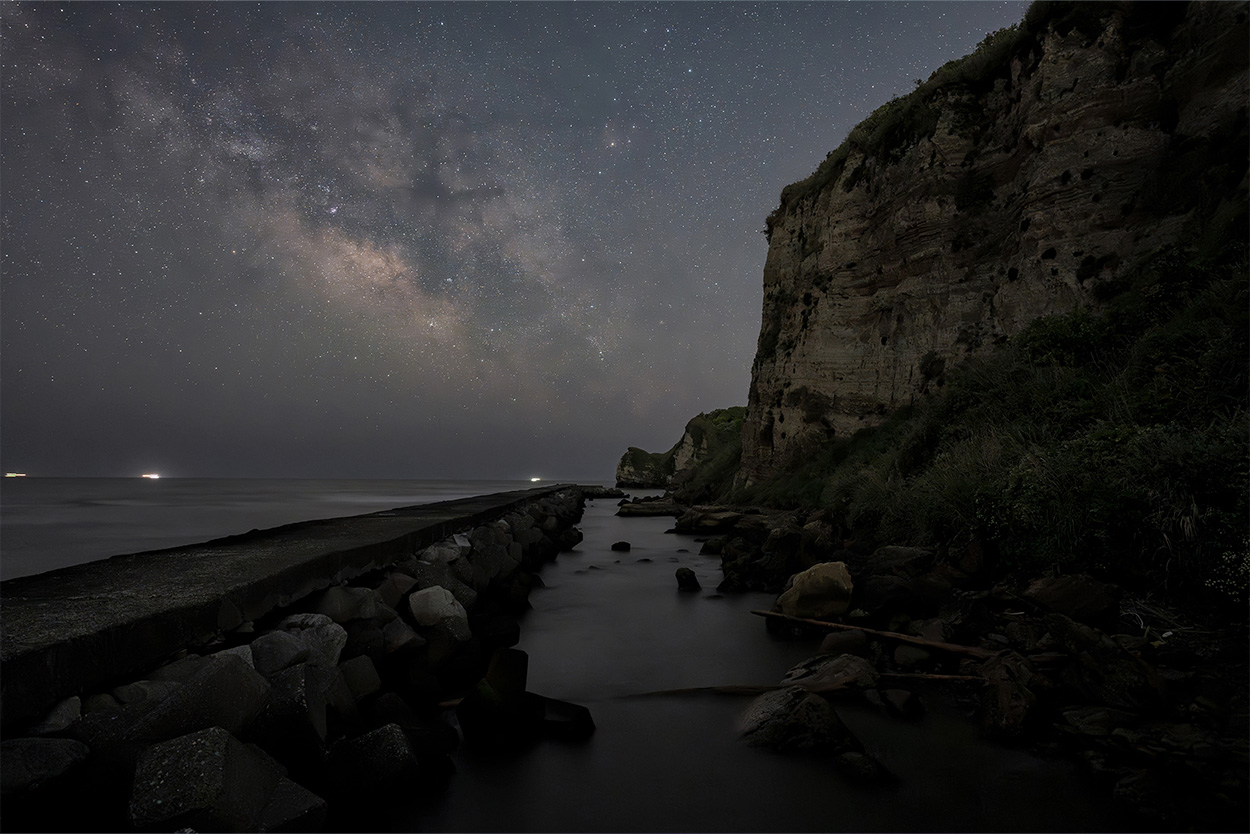 Starry night sky over a rocky coastline with cliffs, a calm sea, and distant lights on the horizon.