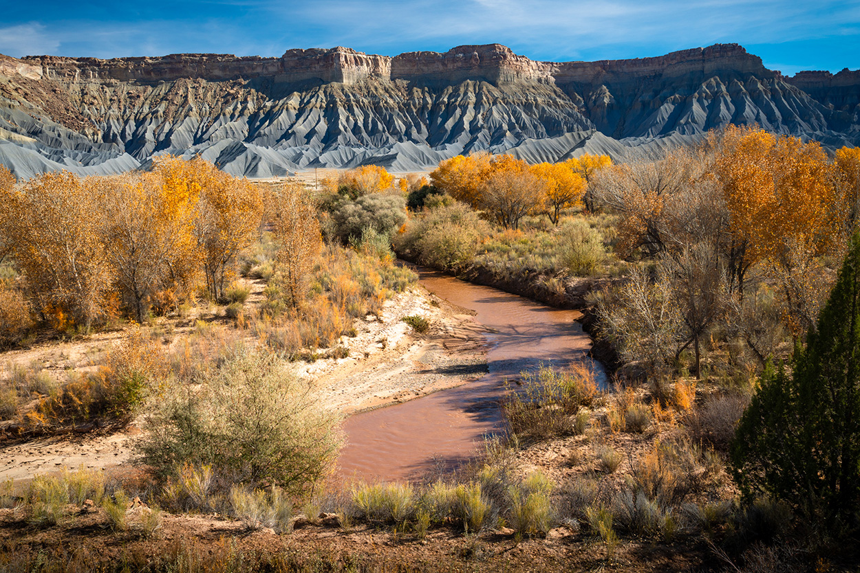 A meandering river flows through a forest of autumn trees, with rugged mountains and layered rock formations in the background under a clear blue sky.