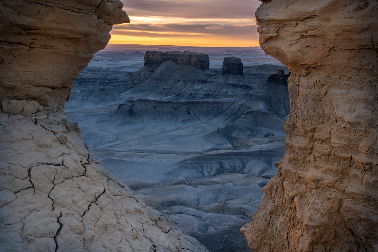 View of a vast desert landscape at sunset, framed by two rugged rock formations, with distant mountains under a colorful sky.