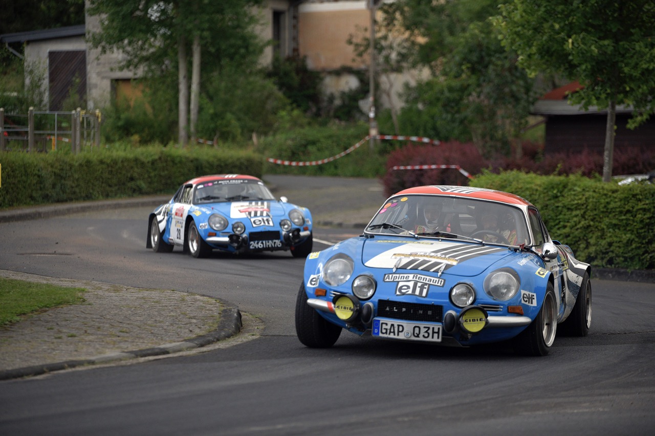 Two vintage blue rally cars with racing decals navigate a curved road lined with greenery and buildings in the background.