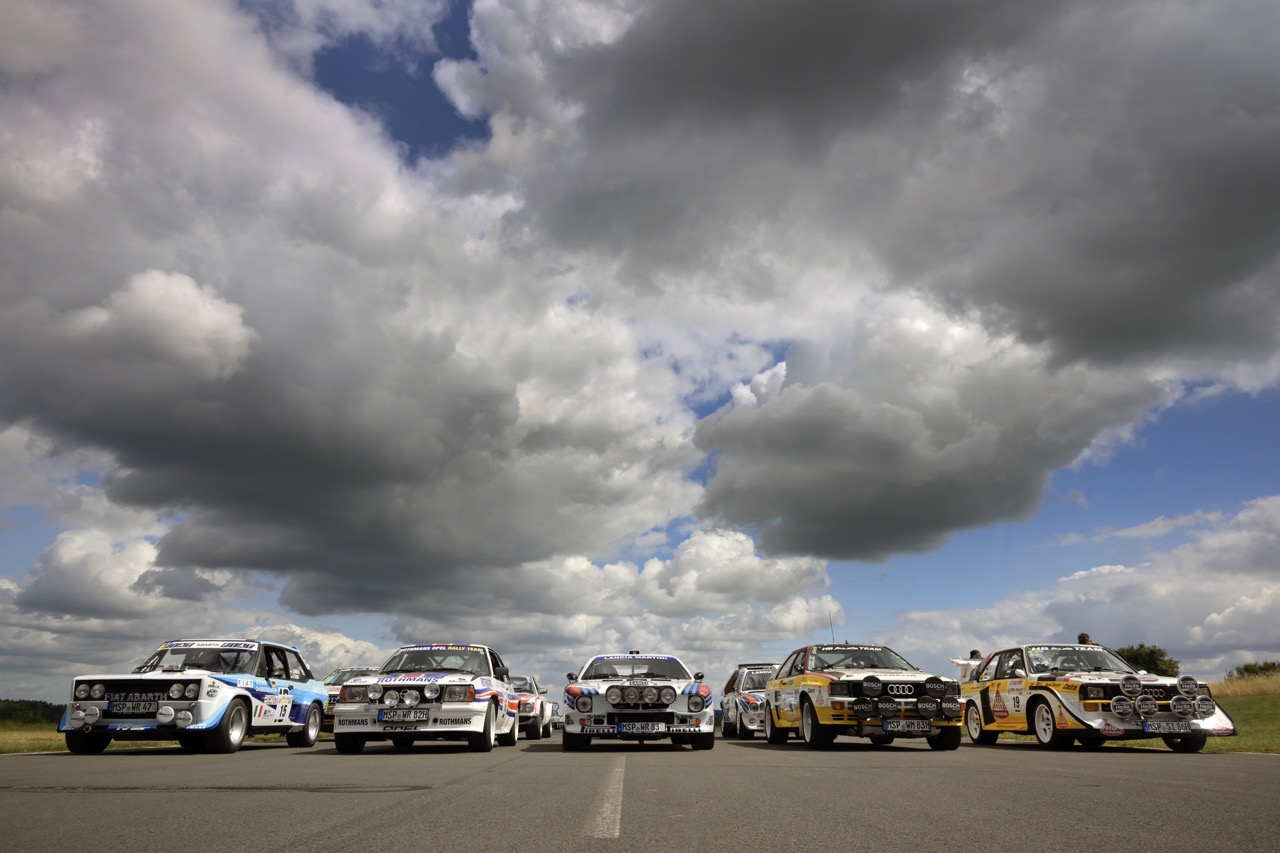 Five vintage rally cars lined up on a road under a dramatic cloudy sky, showcasing various colorful designs and decals.