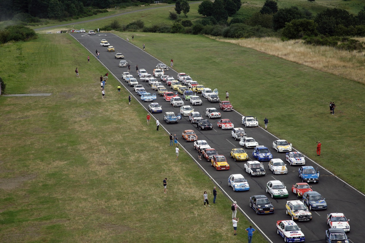 Aerial view of a race track with numerous colorful cars lined up, surrounded by grassy fields and a few spectators walking nearby.