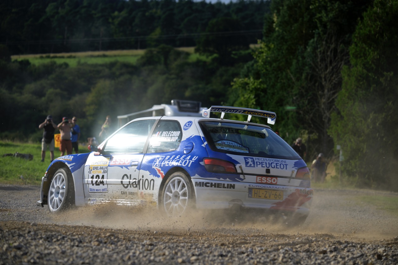 Rally car speeding on a gravel road, kicking up dust, with spectators in the background and lush greenery surrounding the scene.