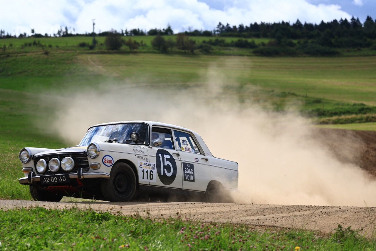A vintage rally car with number 15 races on a dirt track, kicking up dust, set against a backdrop of green fields and a cloudy sky.