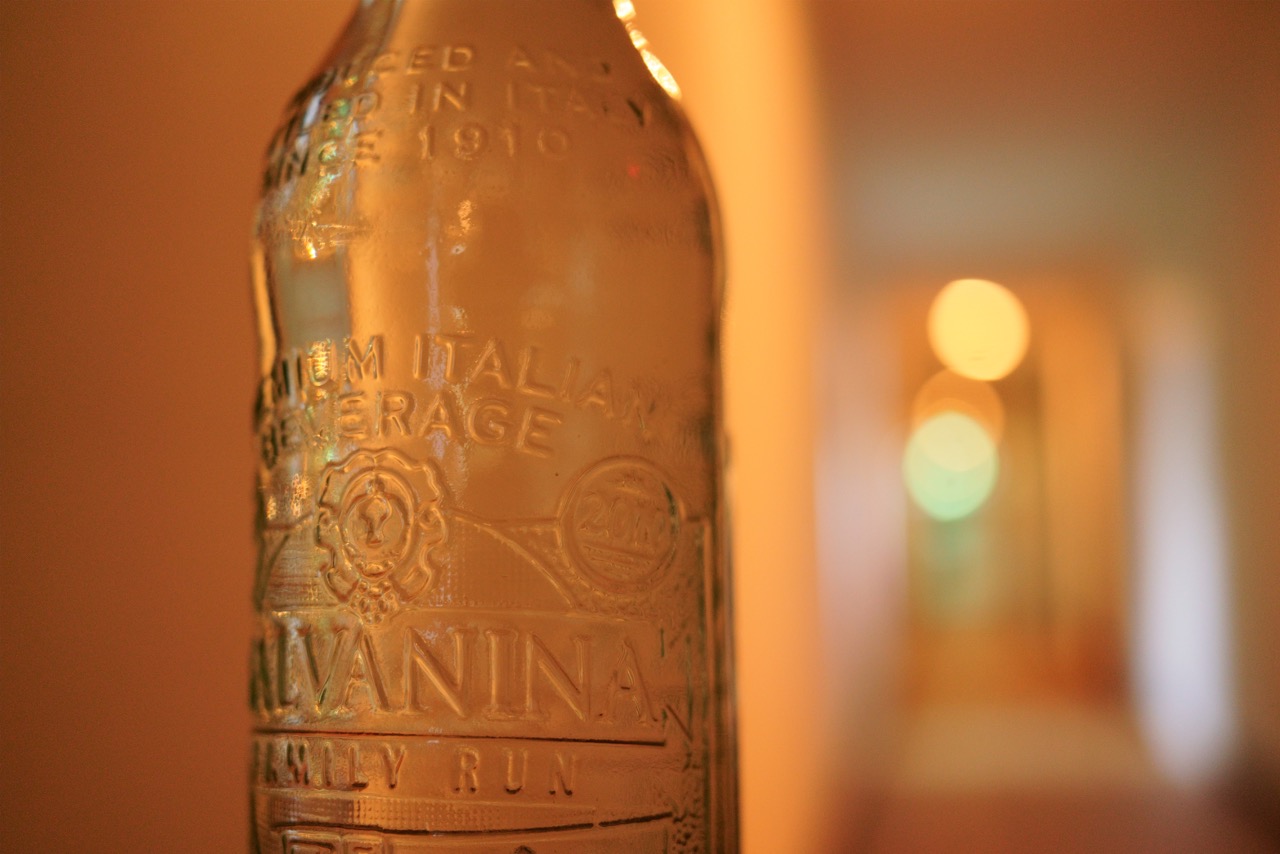 Nahaufnahme einer Vintage-Glasflasche mit geprägter italienischer Beschriftung, vor einem weich unscharfen Hintergrund mit warmem Licht und Bokeh-Effekt.