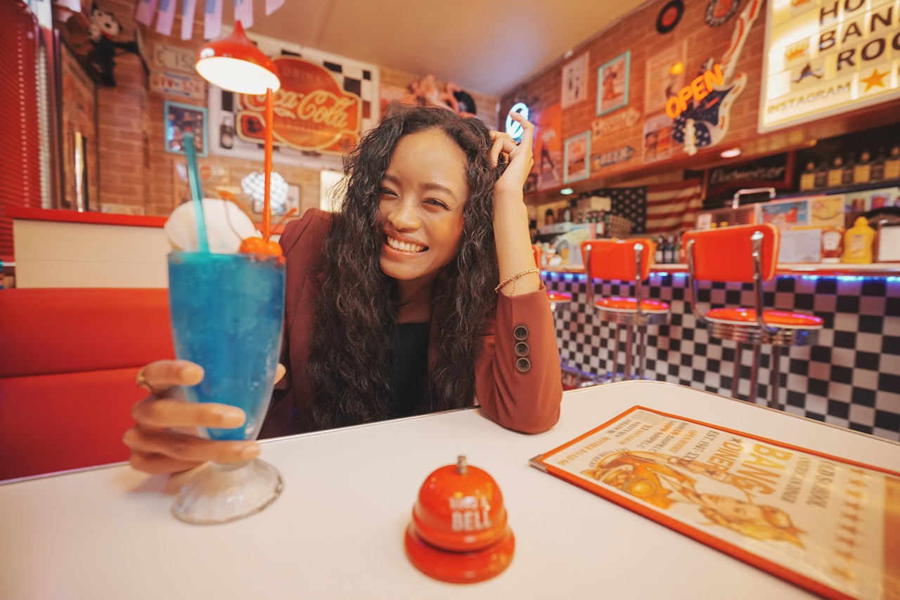 Eine Frau lächelt und hält einen blauen Milkshake in einem Retro-Diner mit roten Sitzbänken und karierter Einrichtung. Auf dem Tisch stehen eine Speisekarte und eine Serviceklingel.