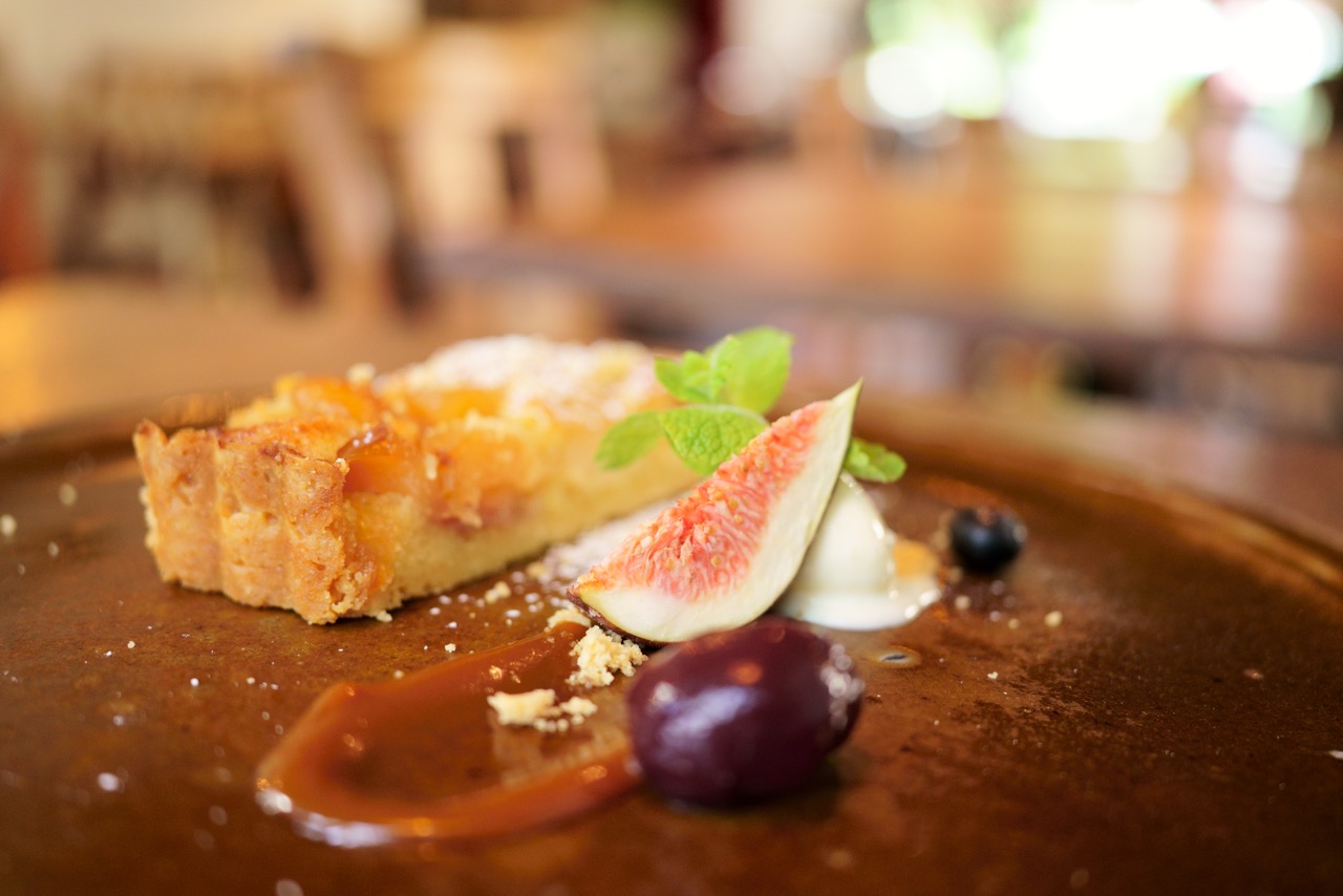 Ein Stück Obsttarte mit Feige, Trauben, Blaubeeren und Minze auf einem braunen Teller, mit Karamellsauce verfeinert, vor unscharfem Hintergrund.