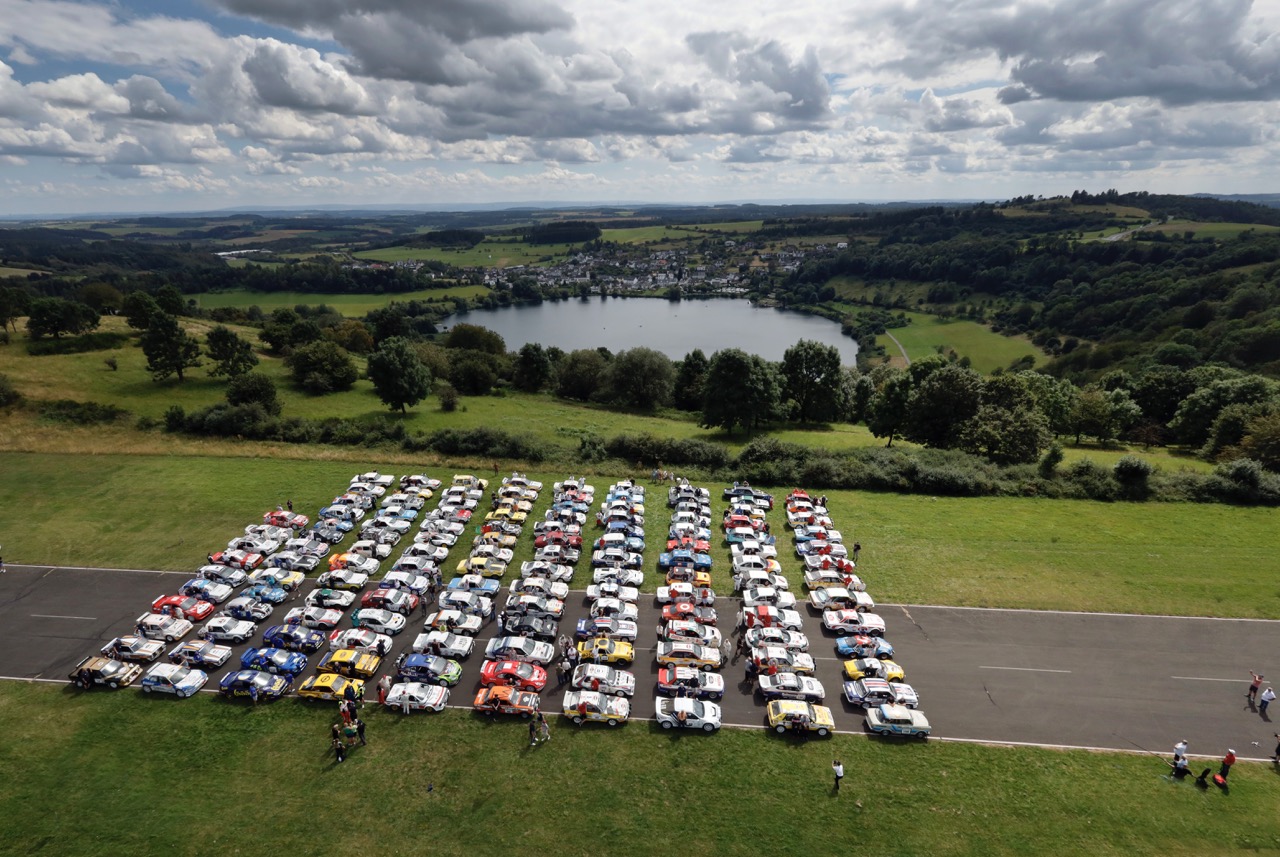Aerial view of numerous classic cars parked in rows on a road, surrounded by lush green landscape and a lake under a cloudy sky.