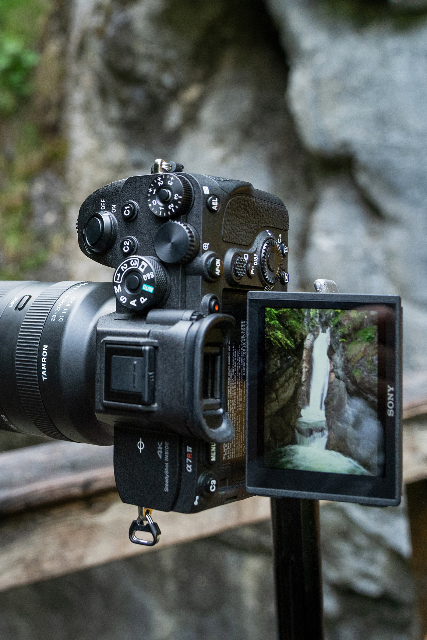 A camera on a tripod capturing a waterfall, displayed on its screen; rocky background and blurred wooden railing in the foreground.