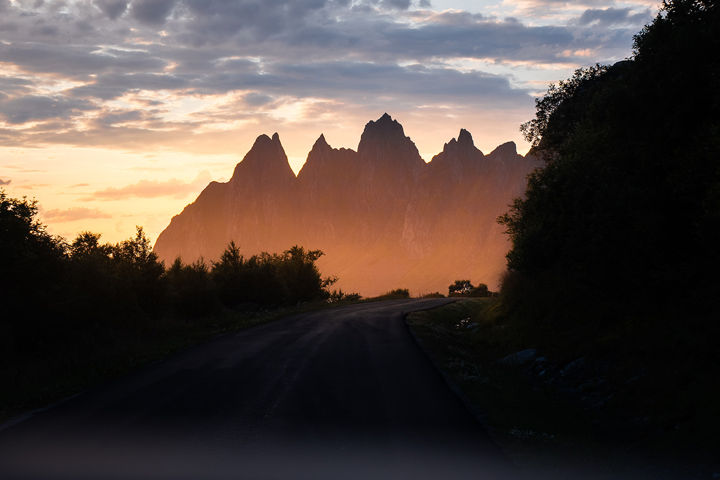 A winding road leads to rugged mountains silhouetted against a glowing sunset sky and lined with dark trees.