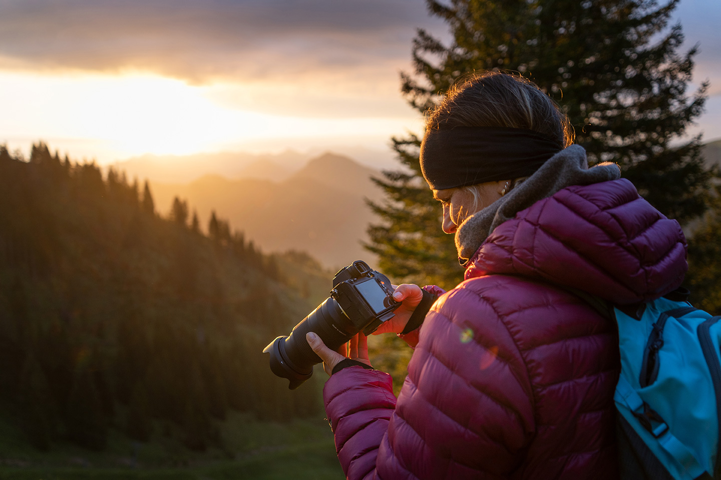 Eine Person in roter Jacke und mit einem Stirnband steht auf einem Berg und betrachtet eine Kamera, während die Sonne gerade in leuchtenden Farben untergeht.
