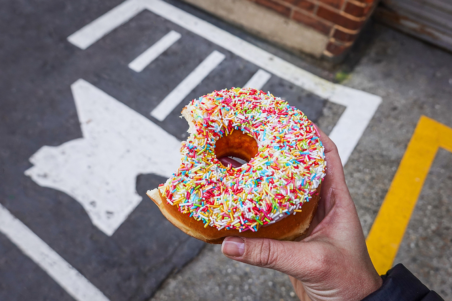 Hand, die einen Donut mit weißem Zuckerguss und bunten Streuseln hält, auf einer Straße mit sichtbarer Straßenmarkierung und einer Backsteinmauer im Hintergrund.