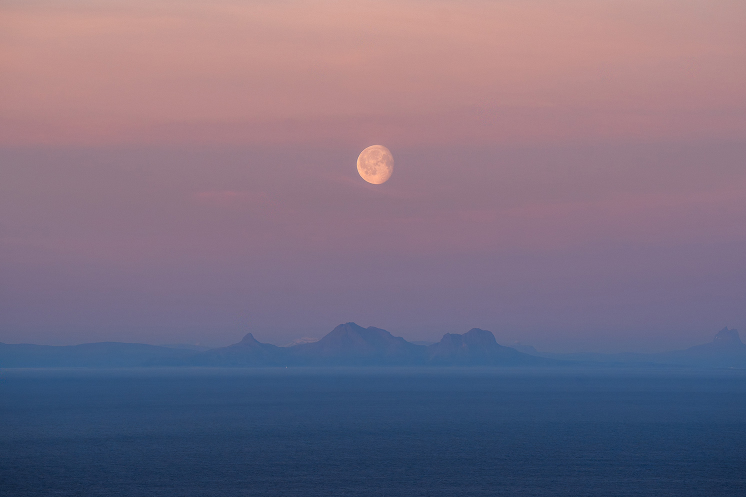 A full moon rises over a calm sea, while in the distance the silhouettes of mountains stand out against a pink and blue evening sky.