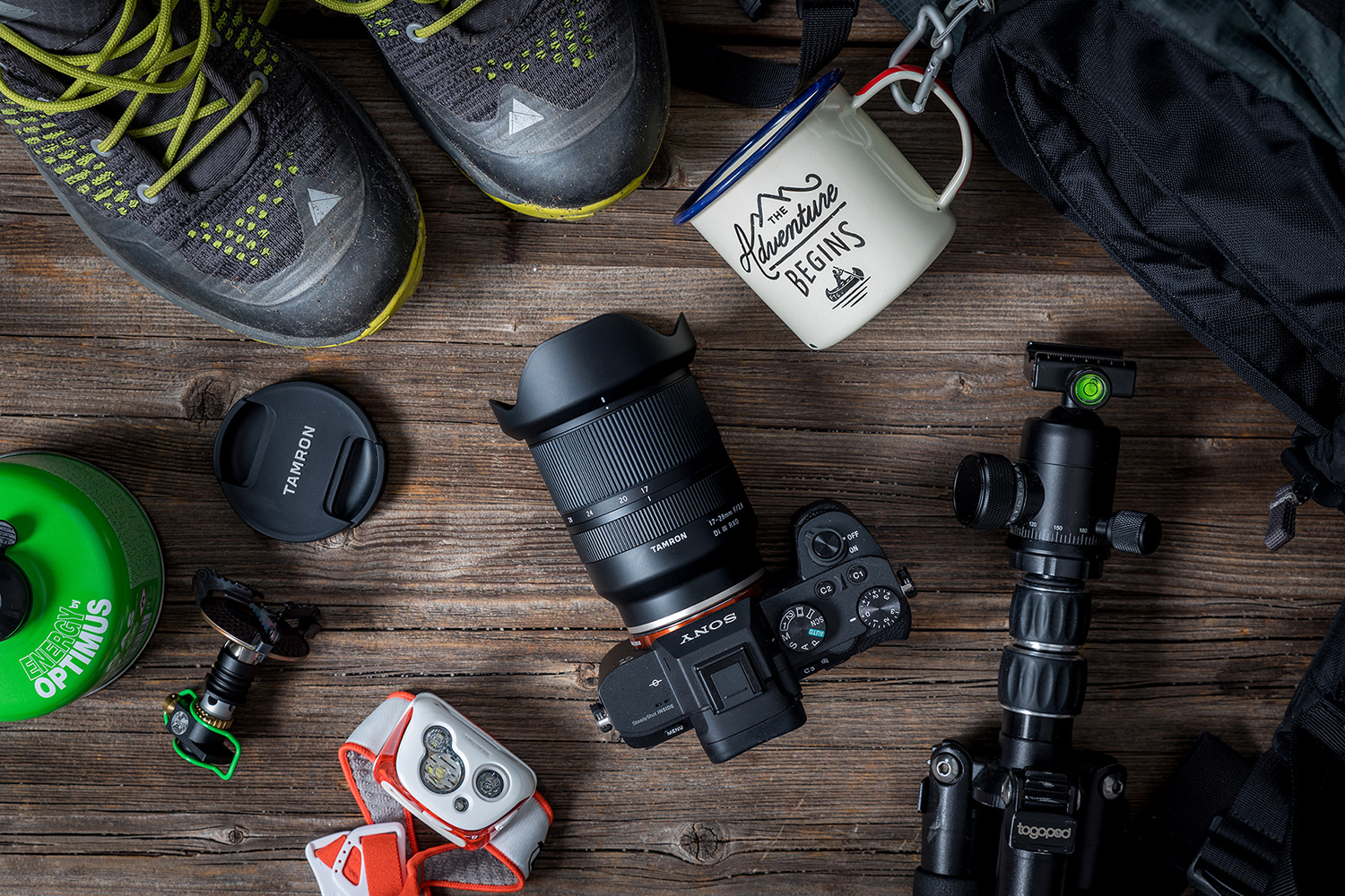 Auf einer Holzfläche liegen eine Kameraausrüstung, ein Stativ, Wanderstiefel, ein Becher, eine Gaskartusche, ein GPS-Gerät und ein Rucksack.