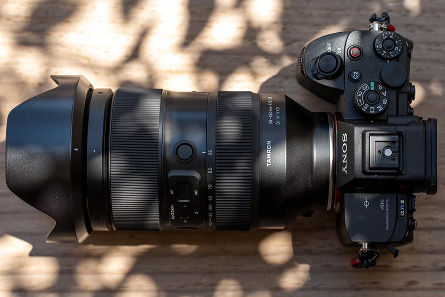 A professional camera with a large lens rests on a wooden surface, partially shaded by leafy shadows.