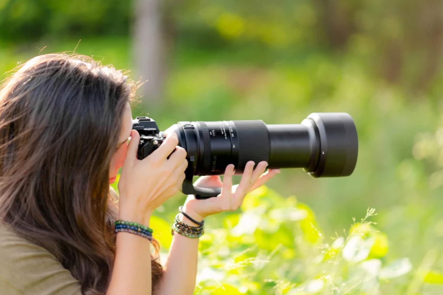Person with long hair using a camera with a large telephoto lens, surrounded by greenery.