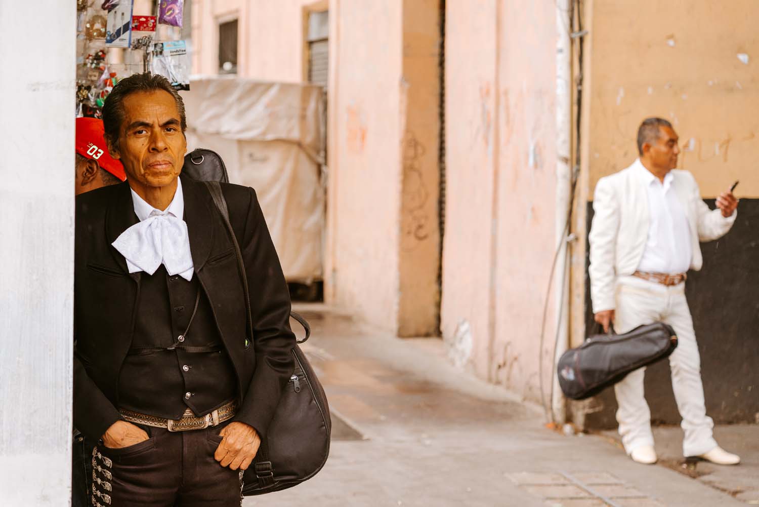 A man in a black mariachi suit leans against a wall, holding a guitar case. Another man in white stands in the background, also with a guitar case.