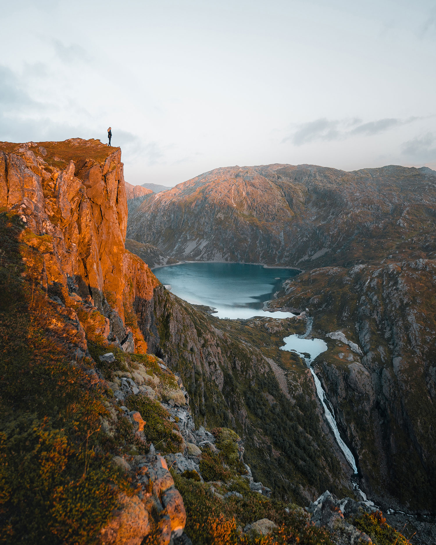 Eine Person steht am Rand einer Klippe und blickt unter klarem Himmel auf eine zerklüftete Berglandschaft mit einem See und einem Bach darunter.