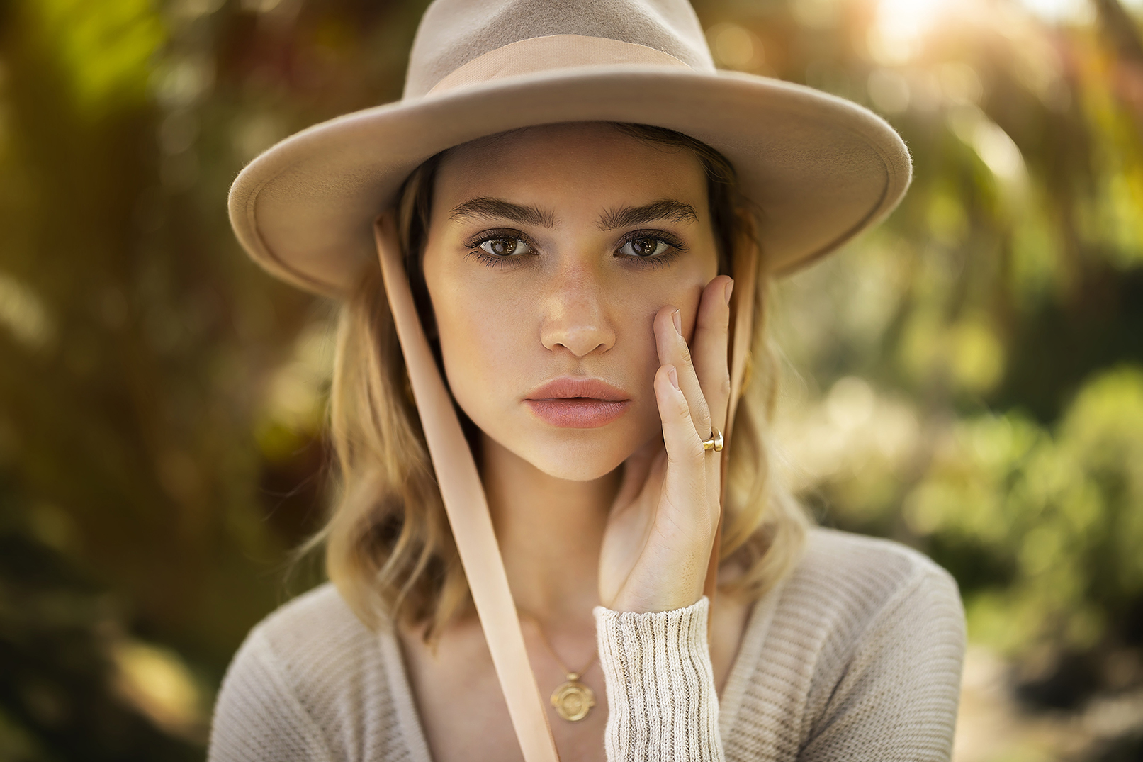 Frau mit einem Hut mit breiter Krempe und neutralen Klamotten, die ihr Gesicht berührt, vor einem verschwommenen natürlichen Hintergrund.