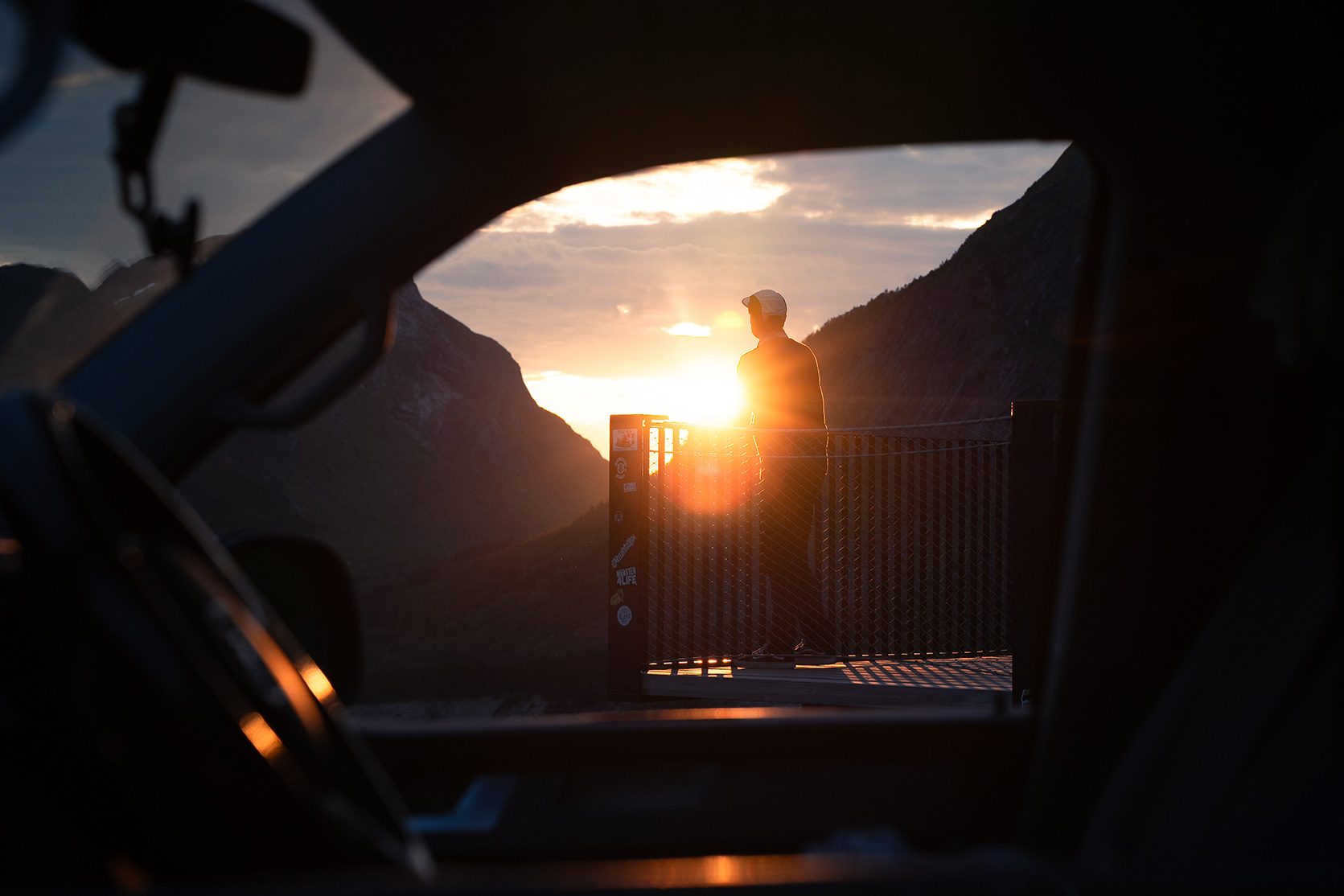 Person standing on a viewing platform at sunset, seen through a car window, with mountains in the background.