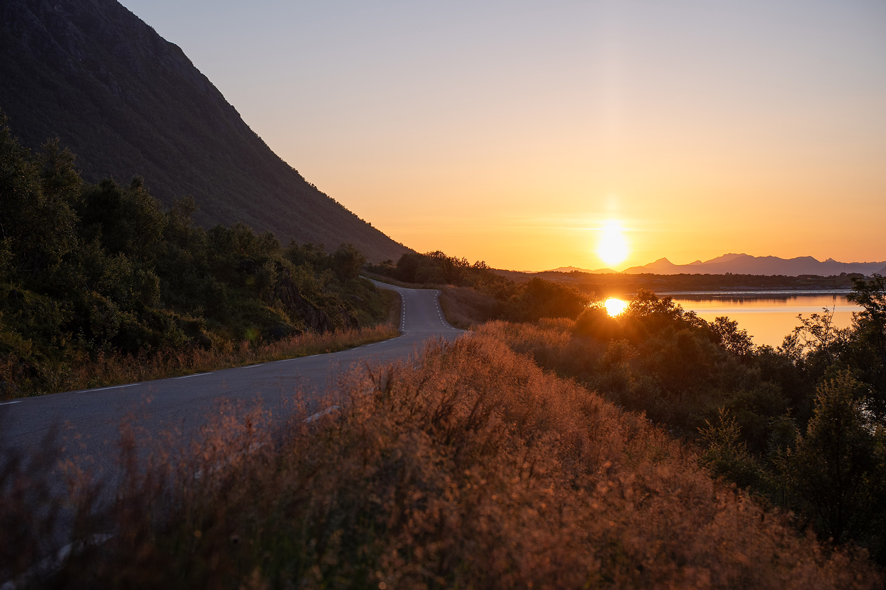 A winding road beside a tranquil lake at sunset, with a mountain silhouette and golden light illuminating the sky and landscape.