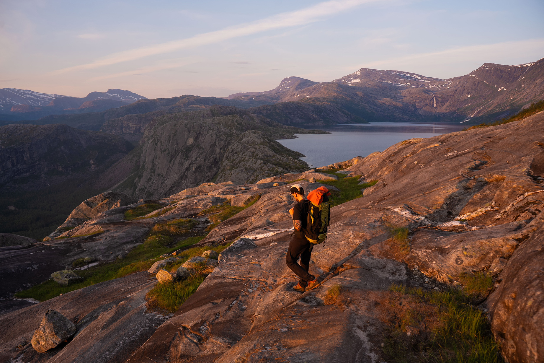 A hiker with a backpack stands on a rocky mountainside at sunset, looking out at a lake and distant mountains under a pink sky.