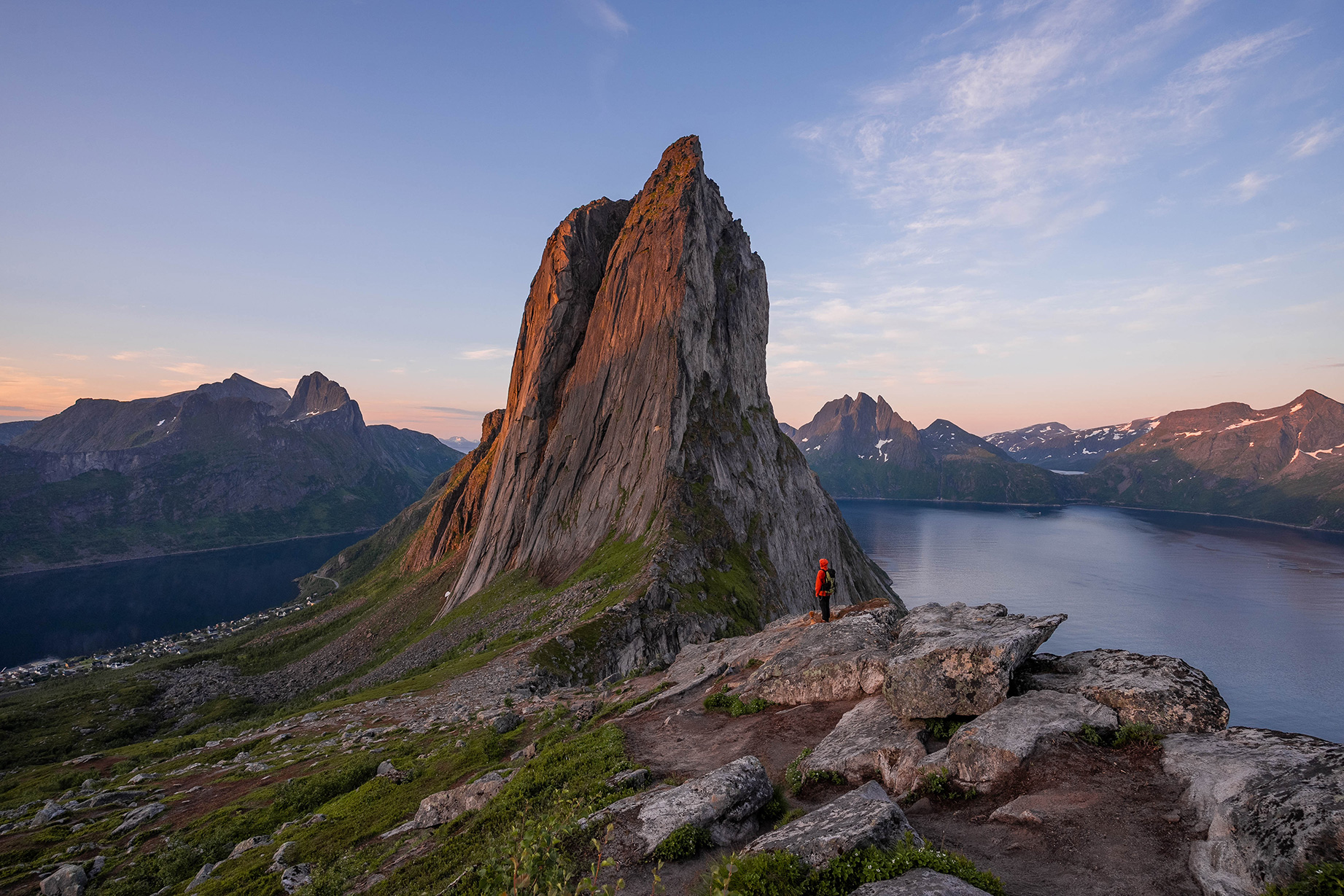 A person stands on rocky terrain and gazes at a dramatic mountain peak and a tranquil fjord at sunset, with snow-capped mountains in the distance.