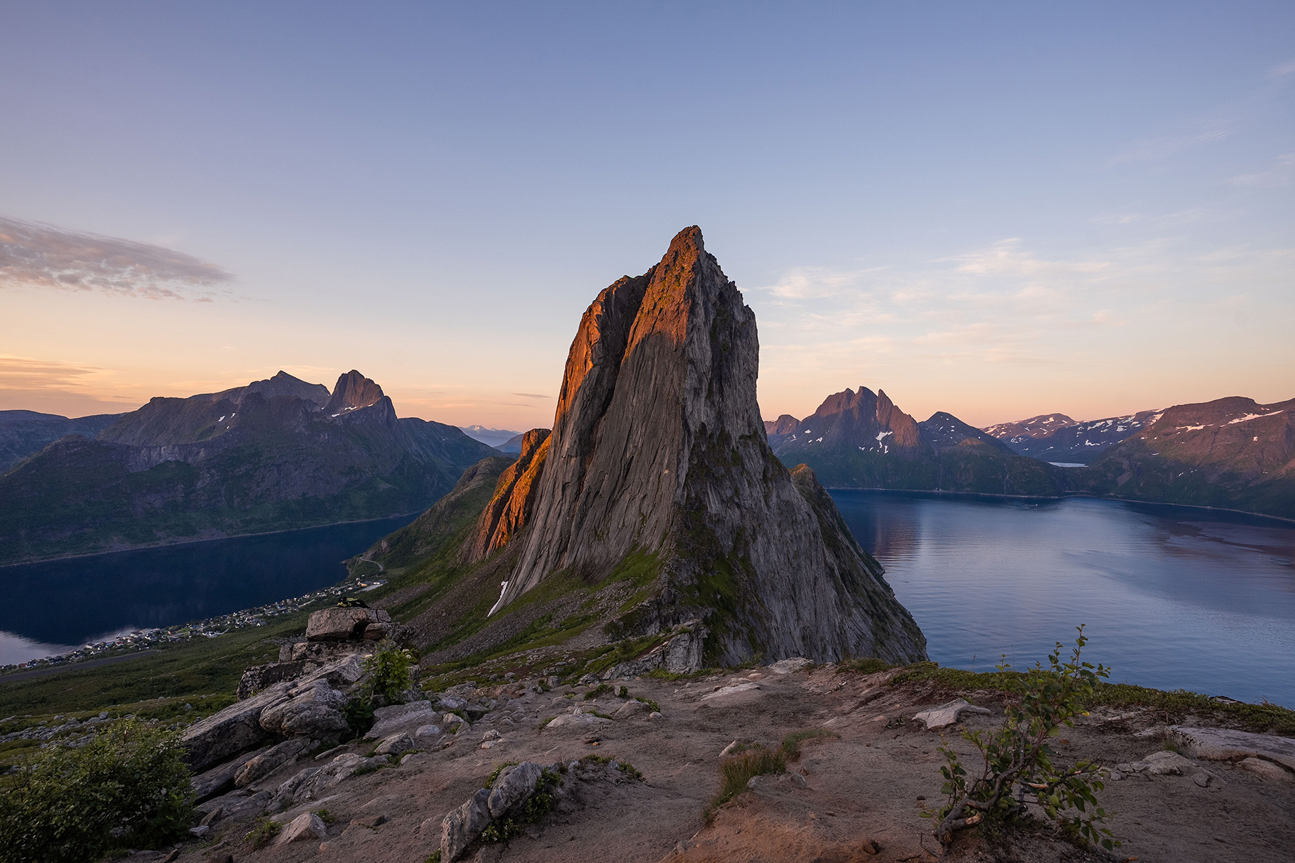A rugged mountain peak at sunset, surrounded by a tranquil lake and distant mountains under a clear sky.