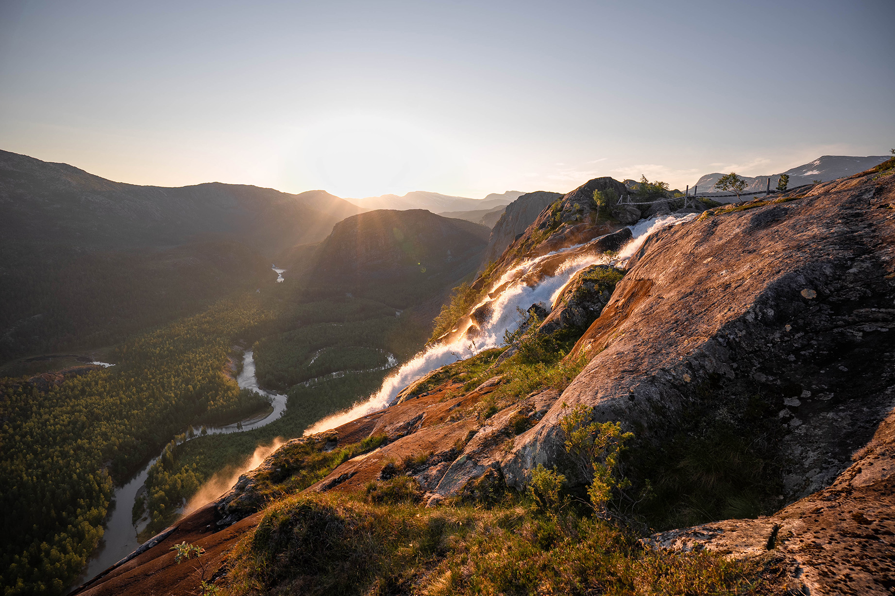 Sunset over a mountain landscape with a winding river and waterfalls cascading over rocky cliffs.