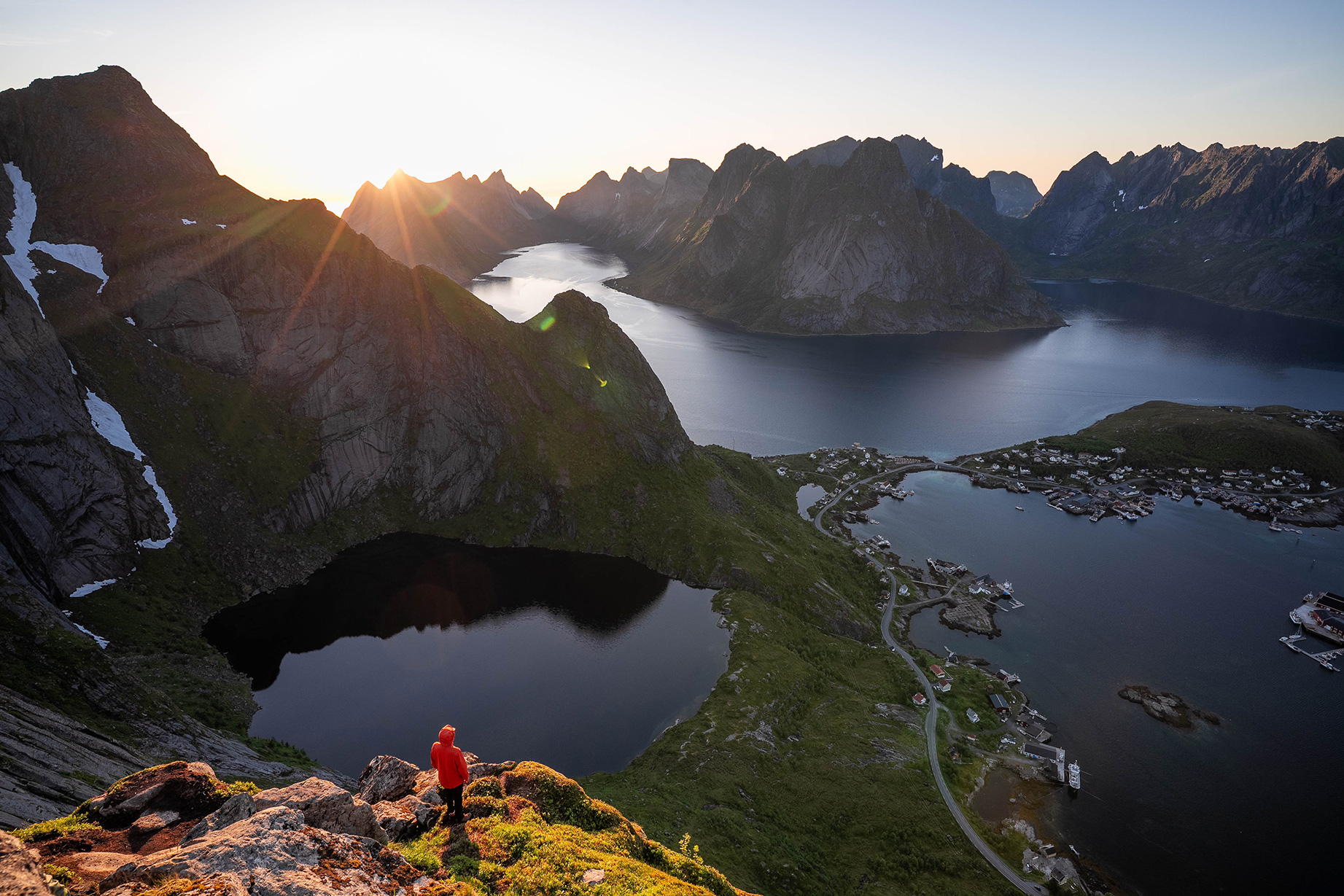 A person wearing a red jacket stands on a cliff and looks out over a picturesque fjord landscape with mountains and the setting sun in the background.