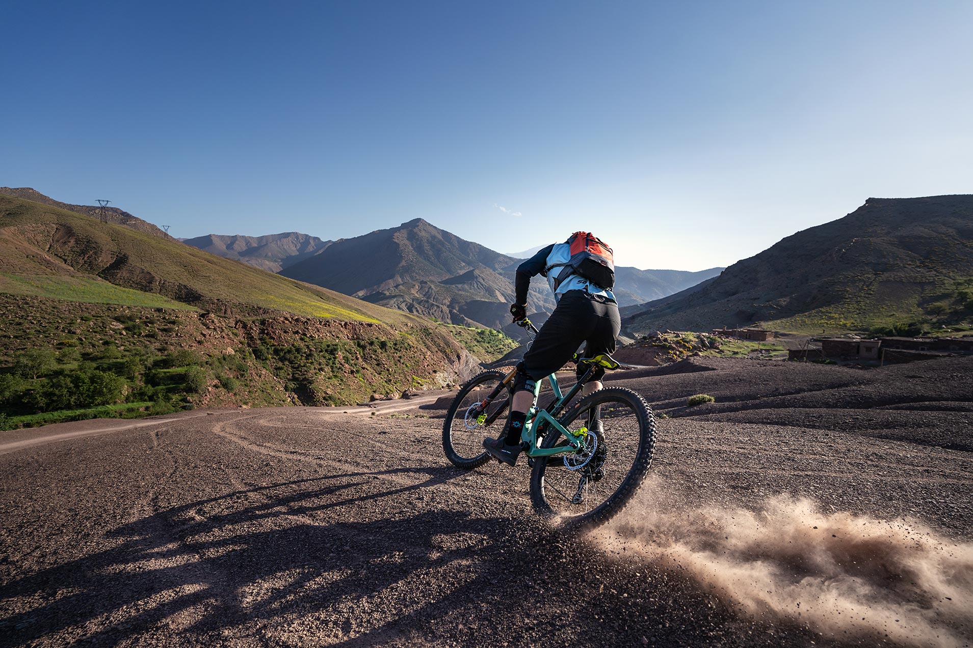 Un ciclista di mountain bike solleva una nuvola di polvere su un sentiero roccioso, sullo sfondo di un vasto paesaggio montano sotto un cielo azzurro e limpido.
