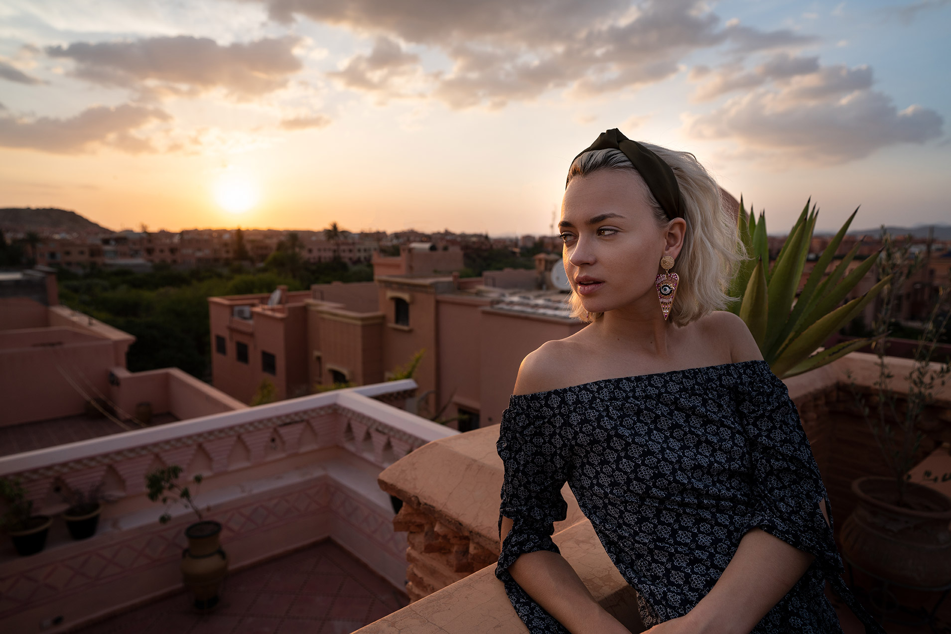 Una donna con un abito senza spalline su un tetto, che ammira il tramonto su un panorama urbano con edifici color terracotta e un cielo nuvoloso.