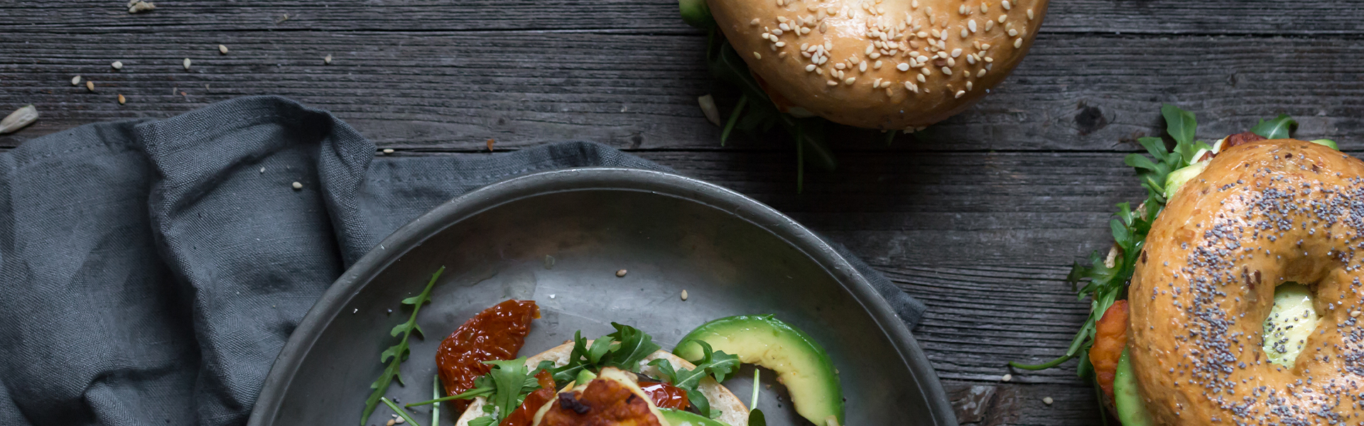 Gegrillter Halloumi, Avocado, sonnengetrocknete Tomaten und Rucola auf einem Teller, dazu Körnerbrötchen auf einem rustikalen Holztisch.