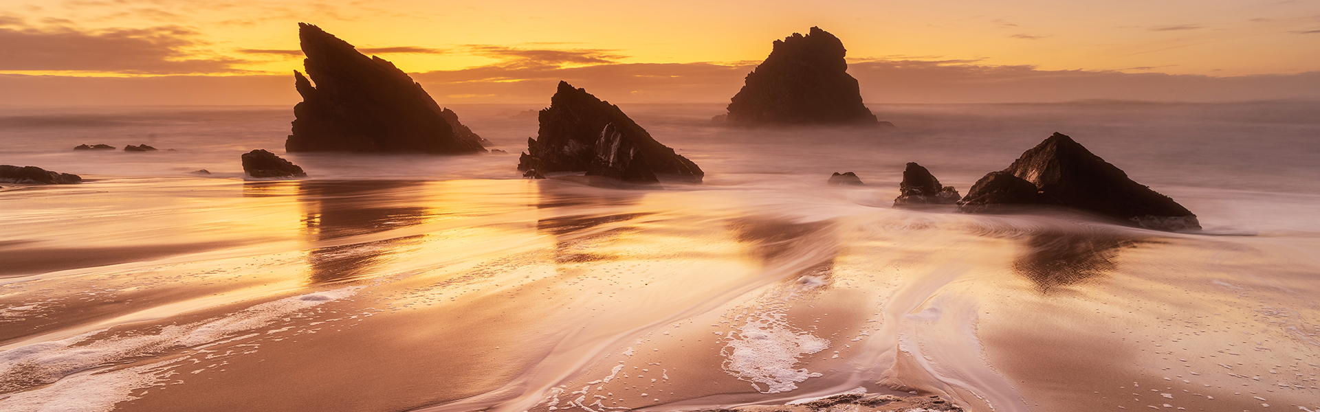 Sonnenuntergang über einem ruhigen Strand mit Felsformationen, der goldene Himmel spiegelt sich im Wasser, und sanfte Wellen zeichnen Muster in den Sand.
