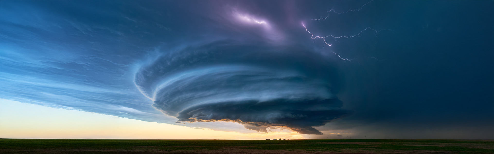 A dramatic supercell thunderstorm with swirling clouds and lightning illuminates the sky over a flat landscape at dusk.