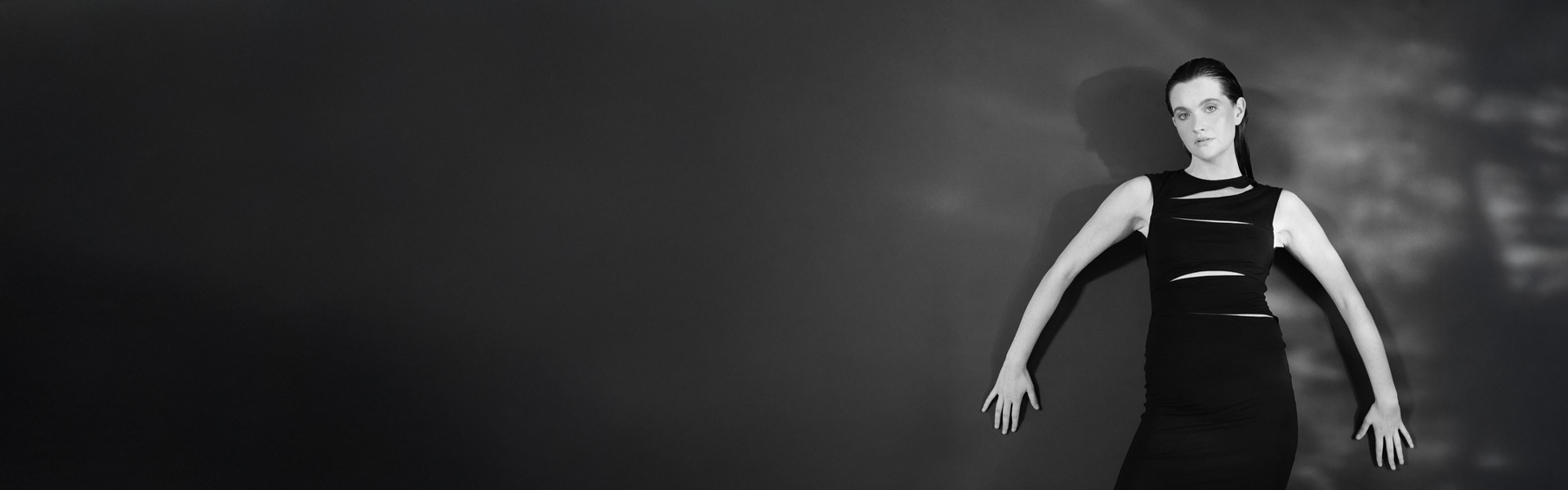 Eine Frau im schwarzen Kleid posiert vor einem dunklen Hintergrund. Das Licht wirft Schatten an die Wand und verleiht der minimalistischen Komposition Tiefe.