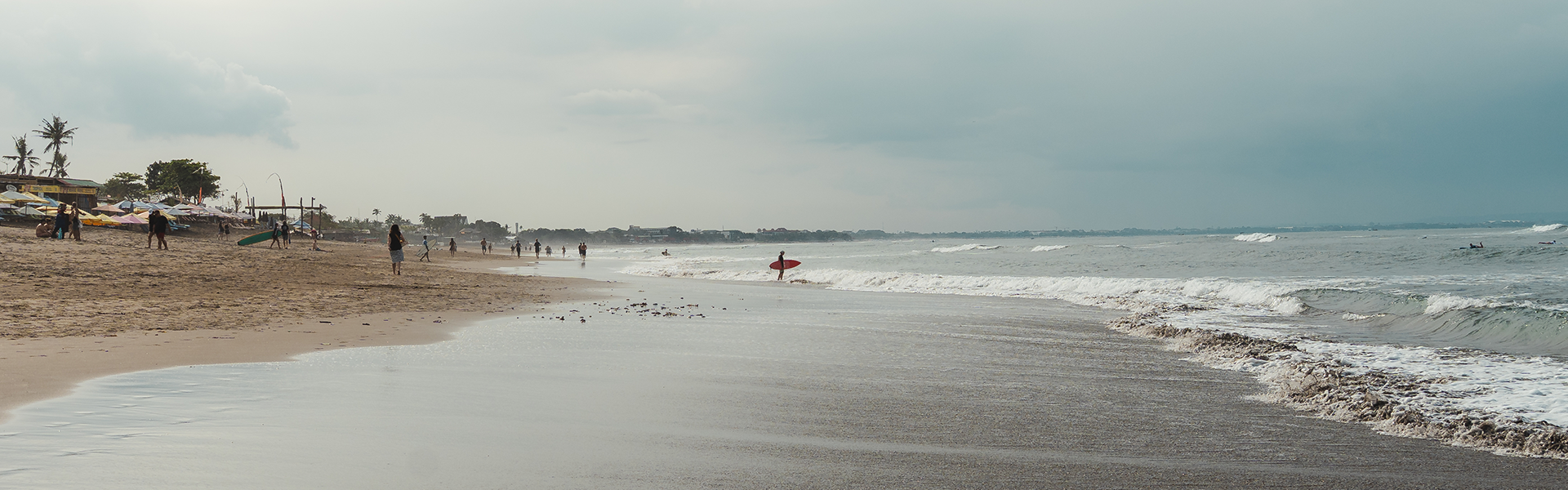Eine wolkige Strandszene mit sanften Wellen, einigen Spaziergängern und einem Surfer, der ein rotes Surfbrett am Ufer entlangträgt.
