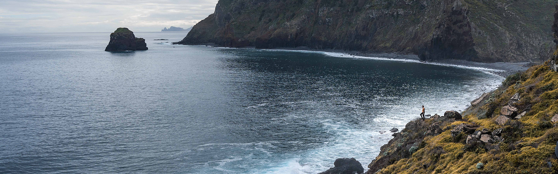 Rocky coastline with waves crashing against cliffs, a lone person stands on a grassy slope, and an overcast sky in the background.